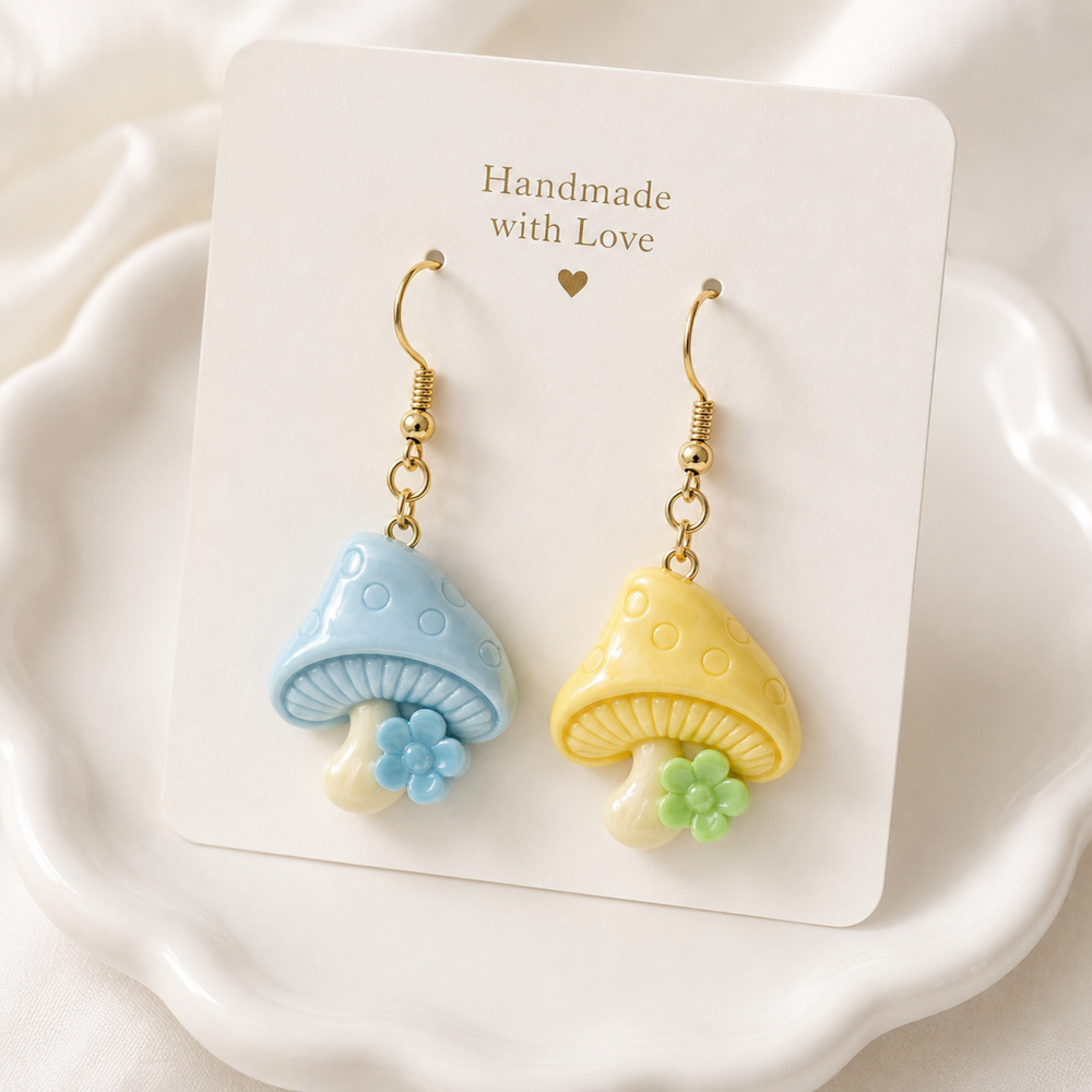 A pair of handmade mushroom-shaped dangle earrings displayed on a white card that reads 'Handmade with Love.' The earrings have gold-tone hooks, one light blue mushroom with a small blue flower and one yellow mushroom with a small green flower, set against a soft white background.