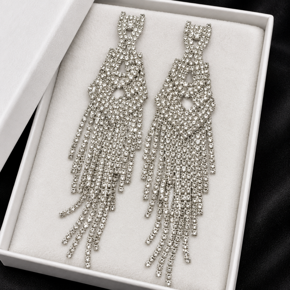 A pair of long, dangling statement earrings covered in clear rhinestones on a white presentation box with a white lining, set against a dark background.