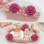 A pink decorative hair comb with glittery rose embellishments, pearls, and rhinestones, shown in a clean studio-style top view and also styled on a mirrored tray with perfume bottles in a lower scene.