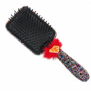 A square hairbrush with a black bristle pad, a handle and frame covered in multicolor sprinkles, a bright red faux-fur accent, and a small orange gingerbread cookie charm, shown on a plain white background.