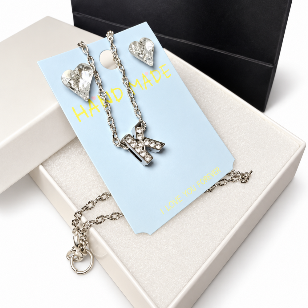 Close-up of a crystal initial K necklace and faceted heart earrings arranged on a blue card inside a white gift box.