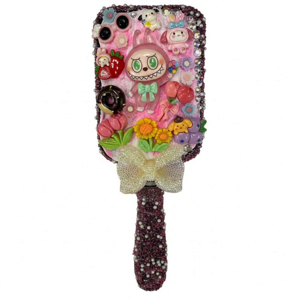 Close up of a decoden paddle brush with a pink monster charm, colorful flowers, fruits, and sparkling crystals