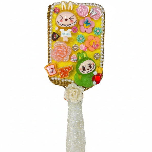 Close up of a handmade white paddle hair brush with a sparkling yellow glitter decoden base and cute monster charms