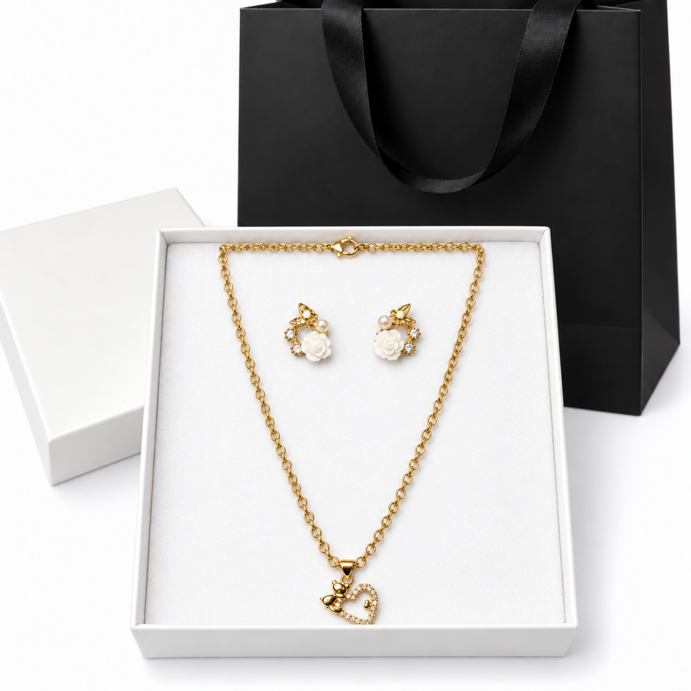 Gold-tone necklace and matching stud earrings displayed in an open white jewelry box, with a black gift bag behind them on a white background.