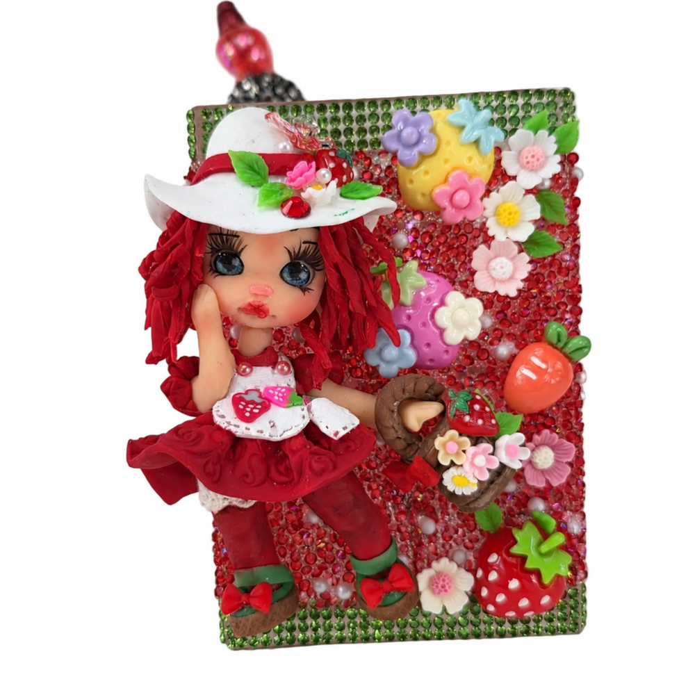 Handmade strawberry cup holder decorated with red rhinestones, green crystal trim, flowers, fruit charms, pearls, and a red-haired doll in a strawberry outfit.
