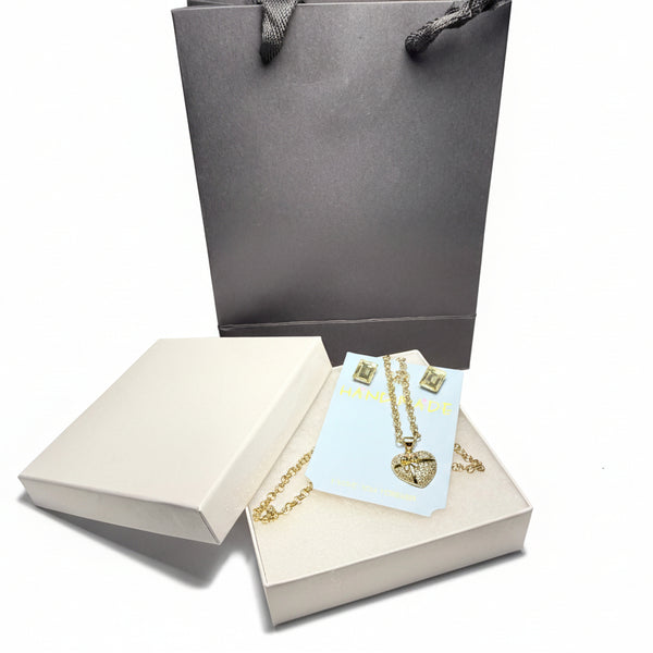 Detailed close-up of a sparkling gold-tone heart pendant necklace and matching earrings, presented in a luxury gift box with a grey gift bag
