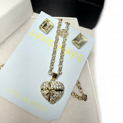 Detailed close-up of a sparkling gold-tone heart pendant necklace and matching earrings, presented in a luxury gift box with a grey gift bag.