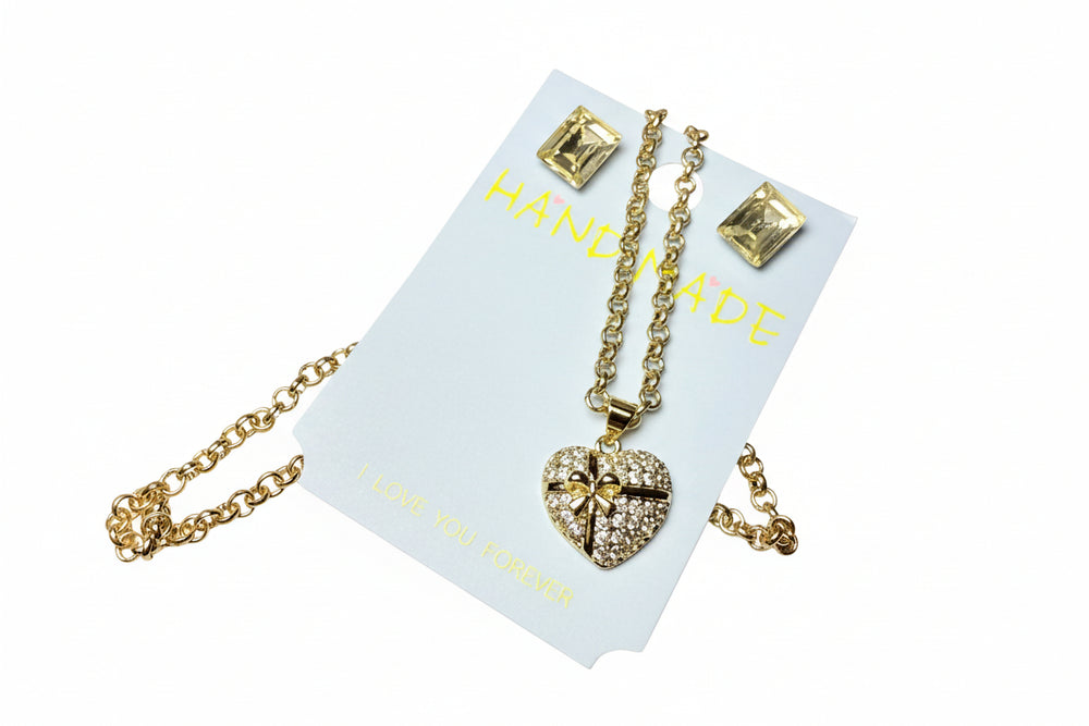 Detailed close-up of a sparkling gold-tone heart pendant necklace and matching earrings, presented in a luxury gift box with a grey gift bag.