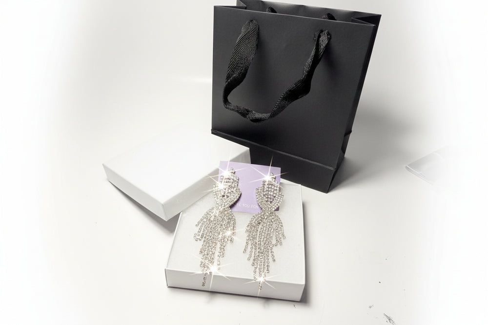 Luxury fringe teardrop crystal earrings displayed on a white gift box next to a black paper bag - elegant gift-ready jewelry presentation.