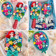 Mermaid-themed gift box with decorated hairbrush, pearl-trimmed hand mirror, mermaid tail earrings, and navy anchor bow with clay doll detail.