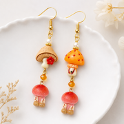 Mushroom resin charms styled as dangling earrings with gold hooks on a white ceramic dish.