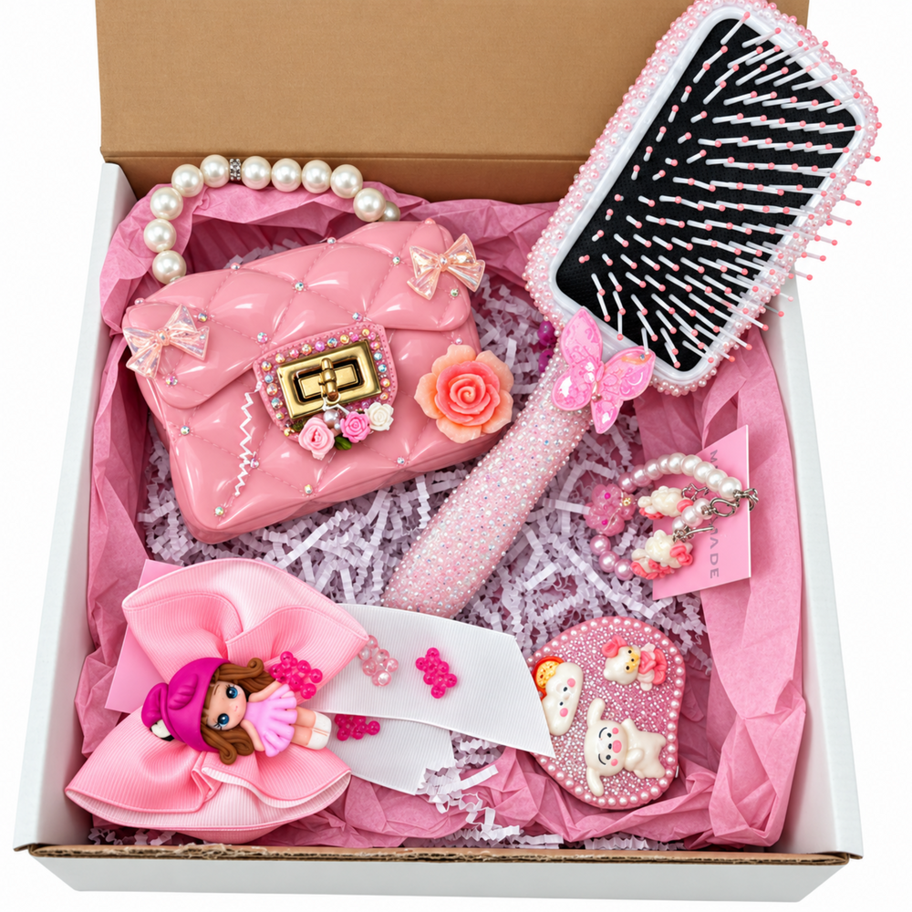 Pink themed gift box featuring a glossy pearl-handle purse, decorated rhinestone hairbrush, oversized pink bow, pearl bracelets, and sparkly accessories arranged in pink tissue paper.