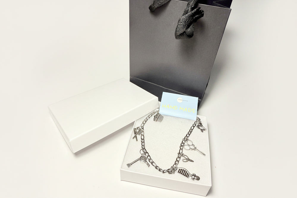Silver charm anklet for hairdressers featuring scissor and comb pendants, displayed on a white gift box with a black luxury gift bag 